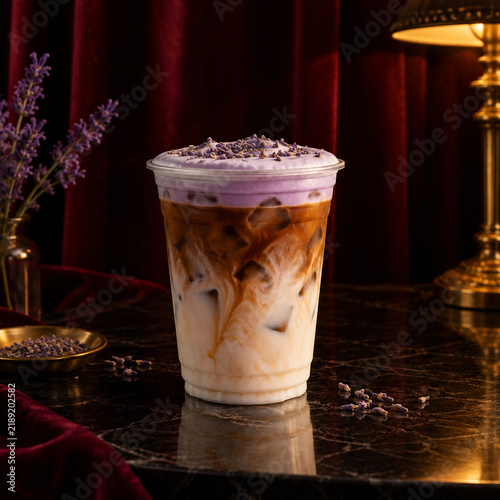 Iced lavender latte with cold foam topping in a plastic cup on a dark marble table with dried lavender flowers and a vintage lamp in a moody setting