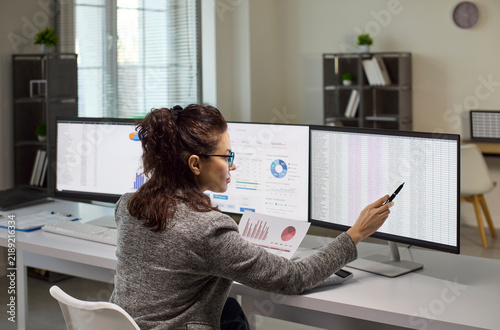 Analyst woman office computer monitors finance data spreadsheet. She reviews a report on dual screens and points at figures to perform audit. Symbolizes precise analysis and financial control.