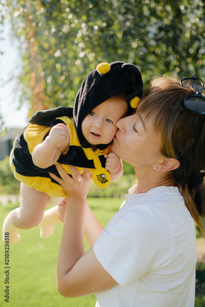 Obraz premium Happy mother kissing cute baby in bee costume outdoors in sunny park, loving family, motherhood and childhood concept.