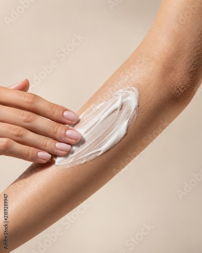 Close-up of a woman applying moisturizing cream to her forearm on a beige background. Luxury skincare routine concept for hydration, body care, dermatology, wellness, and cosmetic advertising.