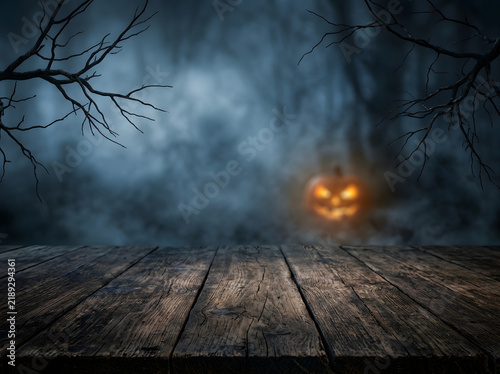 Spooky Halloween background with wooden table | Jack o lantern glowing in foggy forest at night | Rustic wood table surface for product display | Dark misty forest with creepy bare branches