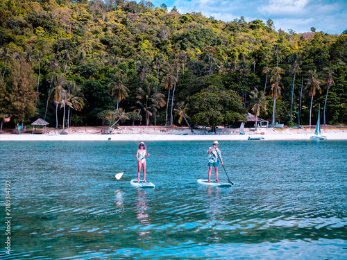 View of couple of tourist are enjoy on water sport paddle sup-board with kayaking along at the edge of the sea beach under crystal clear of water in the island of summer trip