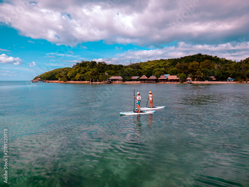 View of couple of tourist are enjoy on water sport paddle sup-board with kayaking along at the edge of the sea beach under crystal clear of water in the island of summer trip