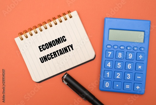 Obraz na plátně A notebook with ECONOMIC UNCERTAINTY text, a blue calculator and a pen against coloured background