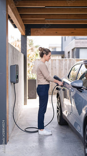 Young woman plugging a charging cable into her grey electric car in a modern home carport, concept of sustainable lifestyle and eco friendly transportation