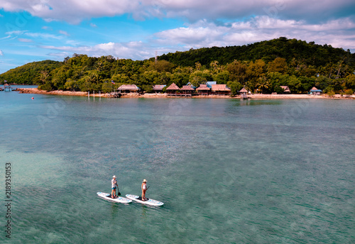 View of couple of tourist are enjoy on water sport paddle sup-board with kayaking along at the edge of the sea beach under crystal clear of water in the island of summer trip