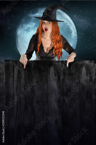 Red-haired witch in black hat peeking from behind dark wooden fence with empty copy space against the backdrop of the moon and night sky. Halloween holiday mockup for text or advertising design