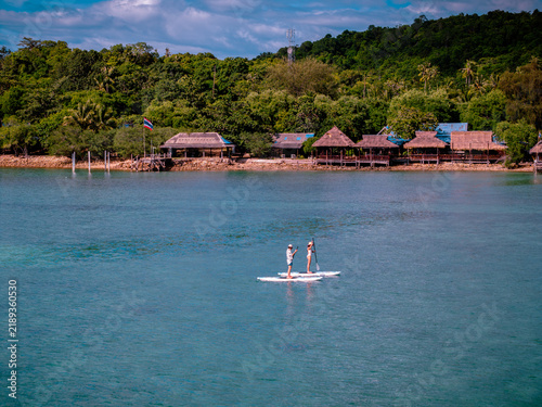 View of couple of tourist are enjoy on water sport paddle sup-board with kayaking along at the edge of the sea beach under crystal clear of water in the island of summer trip