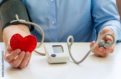 Close view of a person with a blood pressure monitor cuff and pump while holding a red heart, symbolizing cardiovascular health, prevention, care, and diagnosis