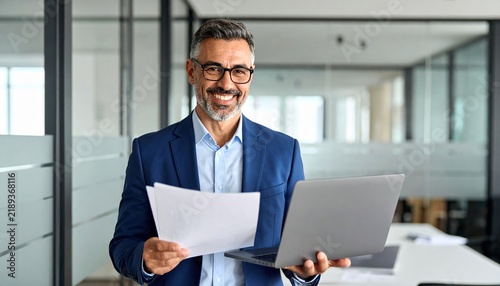 Happy mature businessman holding documents and working on laptop in modern office, professional success