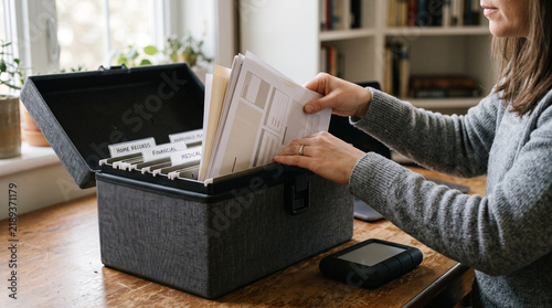 Woman organizing paper documents into a black file box on a wooden desk