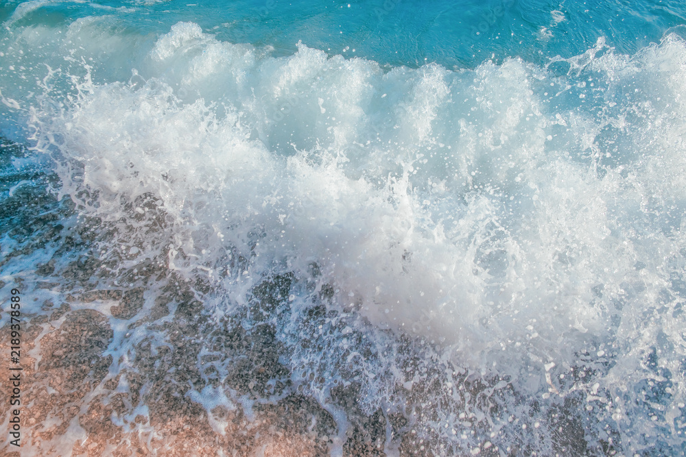 Fototapeta premium Ocean wave crashing onto sandy beach, creating foam and splashes, with clear blue water and sunlight reflecting on the surface in a tropical setting