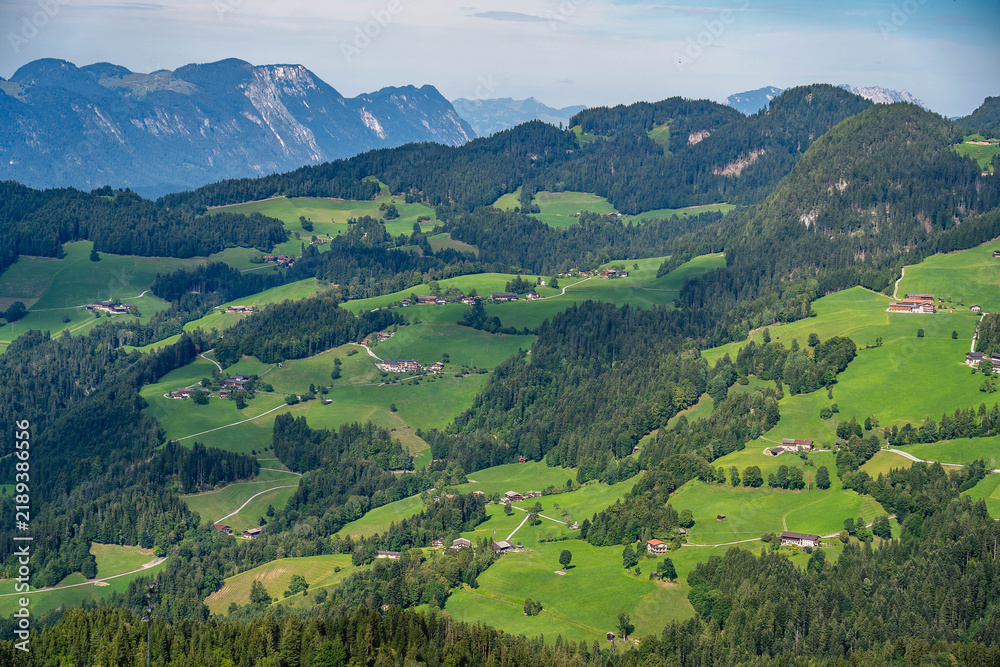 Obraz premium Panoramic view of the Tyrolean Alps in Austria, with rolling green hills, forests, mountain farms and dramatic peaks in the distance.