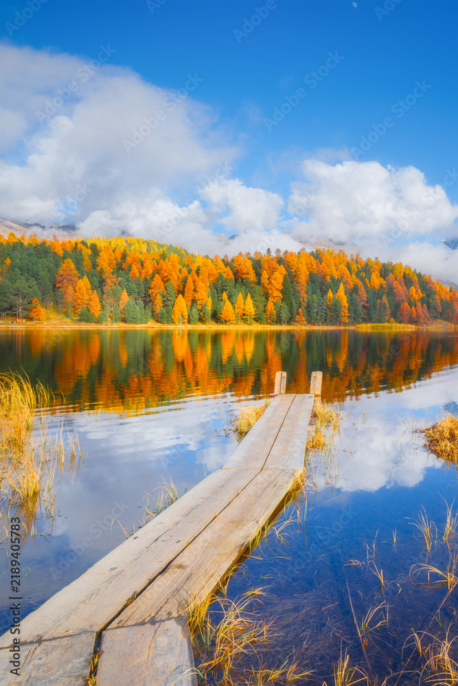 Fototapeta premium Vertical photo. Lake and a small pier. Autumn landscape. Clouds and fall forest. Incredible view of the mountain valley with lake. Background, wallpaper, postcards
