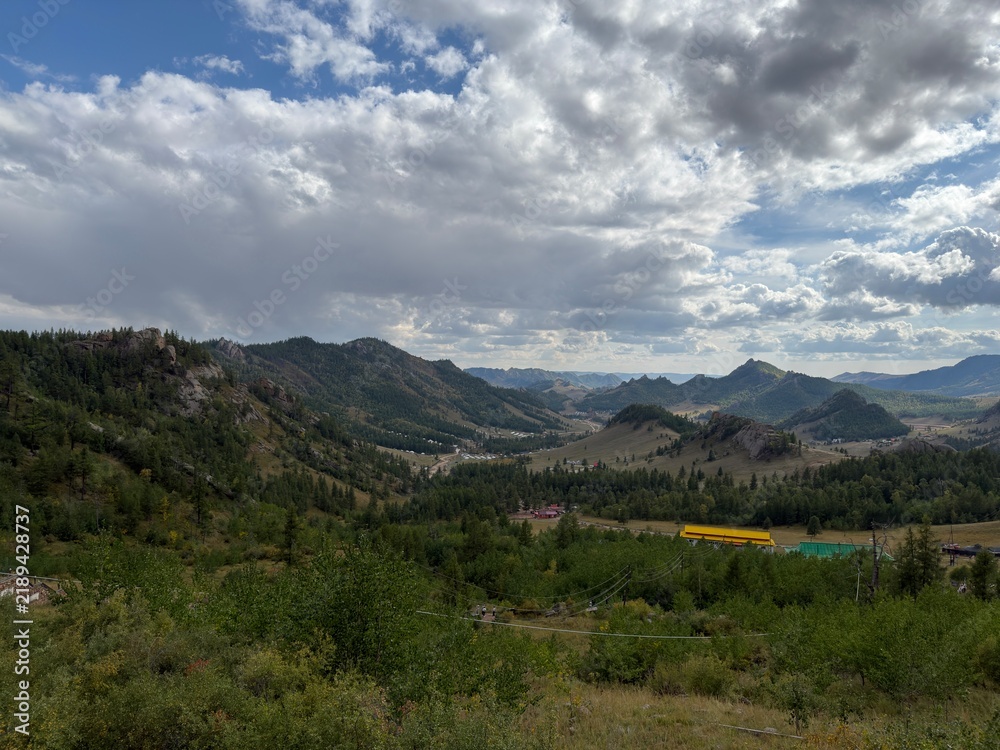 Fototapeta premium Forested Mountain Valley with Rocky Ridges and Scattered Buildings Beneath Dramatic Clouds