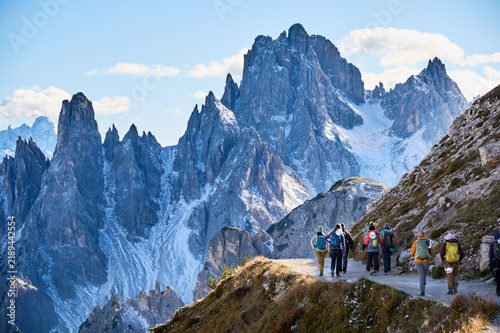 Hiking group follows exposed alpine road beneath towering snow-dusted Dolomites peaks, carrying backpacks and helmets after via ferrata adventure through dramatic high-altitude scenery in Italy