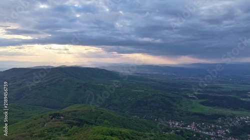 Layered forested mountains and scattered valley settlements fill the landscape during a slow aerial push-in beneath a cloudy evening sky.