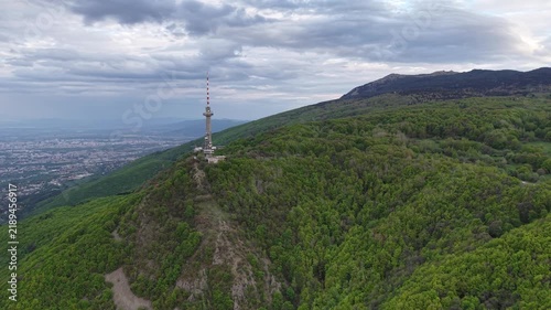 A communications tower rises from a rocky forested mountain ridge above a sprawling city, viewed during a slow aerial glide beneath an overcast sky.