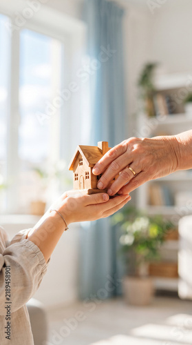 A child s hands hold a wooden house  passed to an adult s hands  symbolizing family  home  and legacy transfer.