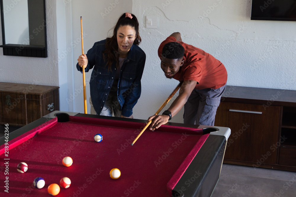 Obraz premium Diverse friends playing on red-felt pool table at home, woman wearing denim jacket, man aiming shot