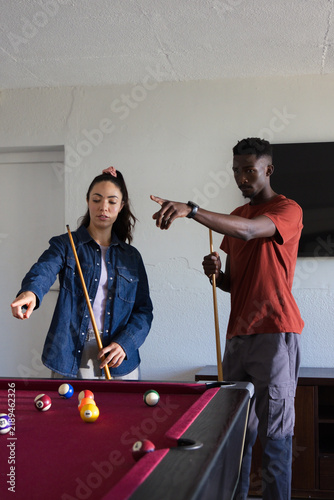 Diverse couple standing at red felt pool table, pointing and planning shots with cues, denim jacket