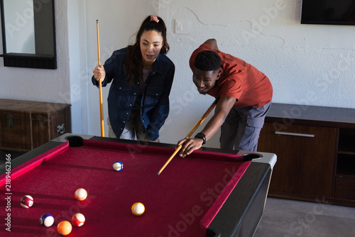 Diverse friends playing on red-felt pool table at home, woman wearing denim jacket, man aiming shot