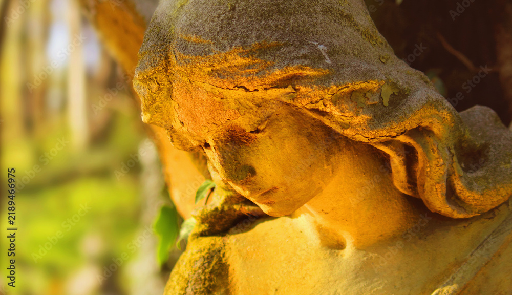 Fototapeta premium Sorrowful angel statue with bowed head illuminated by warm golden sunlight, symbol of grief, remembrance, loss and eternal memory.