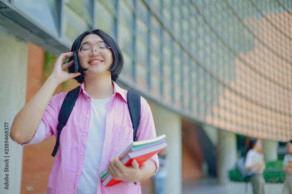 Fototapeta premium Happy asian LGBTQ female student stands in front university building, carrying a backpack and textbooks, while talking on the phone with a university friend who is waiting in the classroom.