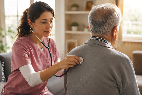 A healthcare worker examines an elderly man with a stethoscope in a domestic setting