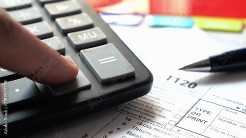Corporate tax accountant finger pressing equal button on calculator over internal revenue service form 1120 with pen