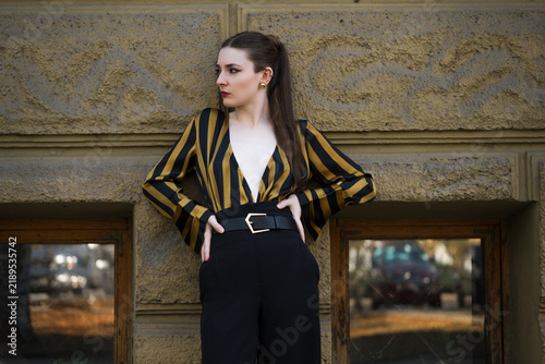 Portrait of an attractive young caucasian woman wearing a yellow and black striped blouse near a building. Fashion, style, beauty.