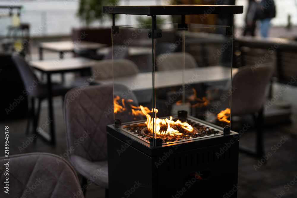 Fototapeta premium Street cozy terrace with comfortable seating places and gas heater in glass box for guests, modern restaurant outdoor patio with couches, tables and fireplace inside glass cabinet, dining lounge zone.