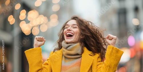 Excited woman celebrates successful shopping trip with bags and yellow coat on street lights background