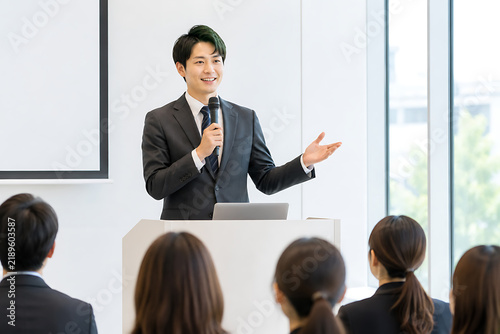マイクを持って明るい表情としぐさで講演する男性ビジネスパーソンのシーン素材