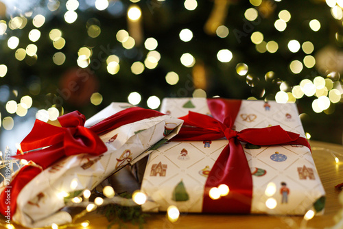 Christmas wrapped gifts tied with red ribbons on a wooden table illuminated by fairy lights, festive concept of gift giving. Holiday present wrapping, cozy Christmas Eve celebration.