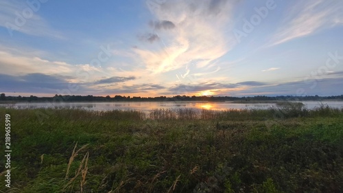 Fotografía Serene lakeside dawn vista showing glowing cloudscape reflected in still waters beyond grassy shores and wild reed beds