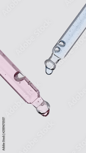 Two glass cosmetic pipettes with pink serum and clear liquid converging tips diagonal composition