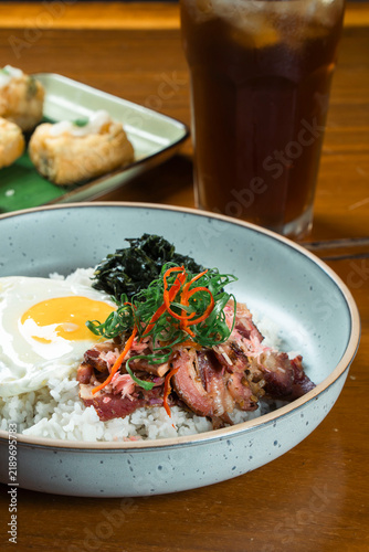 Hearty Breakfast Bowl: A delectable rice bowl topped with a fried egg, savory meat, and fresh garnishes, served alongside a refreshing iced drink, offering a satisfying start to the day.