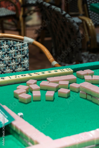 Mahjong Tiles on Green Felt: A close-up view of pink Mahjong tiles scattered across a green felt table, set against a backdrop of wicker chairs, hinting at an engaging game ready to begin.