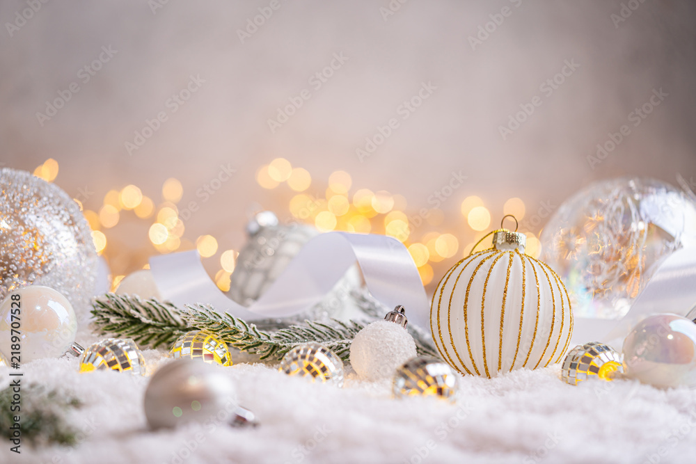 Naklejka premium A Christmas background featuring textured white and gold baubles alongside tiny mirror disco balls and frosted pine on snow, softly lit by warm twinkling lights with copy space.