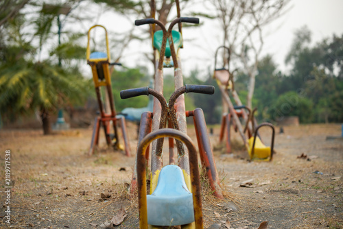 A rusty abandoned seesaw with yellow seats sits on an empty outdoor playground surrounded by dry grass and trees, suggesting nostalgia, neglect and quiet urban decay.