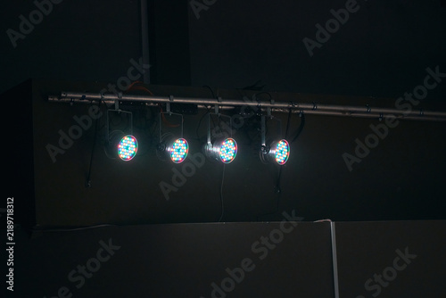 Row of four illuminated stage lights on metal rigging bar in dimly lit room