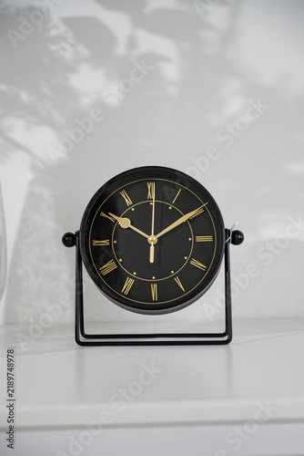 Black analog clock with Roman numerals on the table in the room.