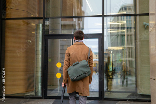 Man in brown coat with bag standing in front of glass office building entrance. Professional commuter arriving at workplace. Modern city lifestyle and urban business environment concept.