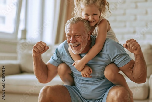 Joyful grandfather giving his smiling granddaughter a piggyback ride while playing in the living room