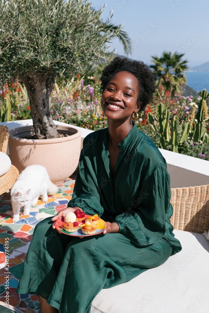 Fototapeta premium Smiling woman in a green dress holding a plate of fresh fruit on a terrace garden with cat and ocean view for summer lifestyle banner and wellness design