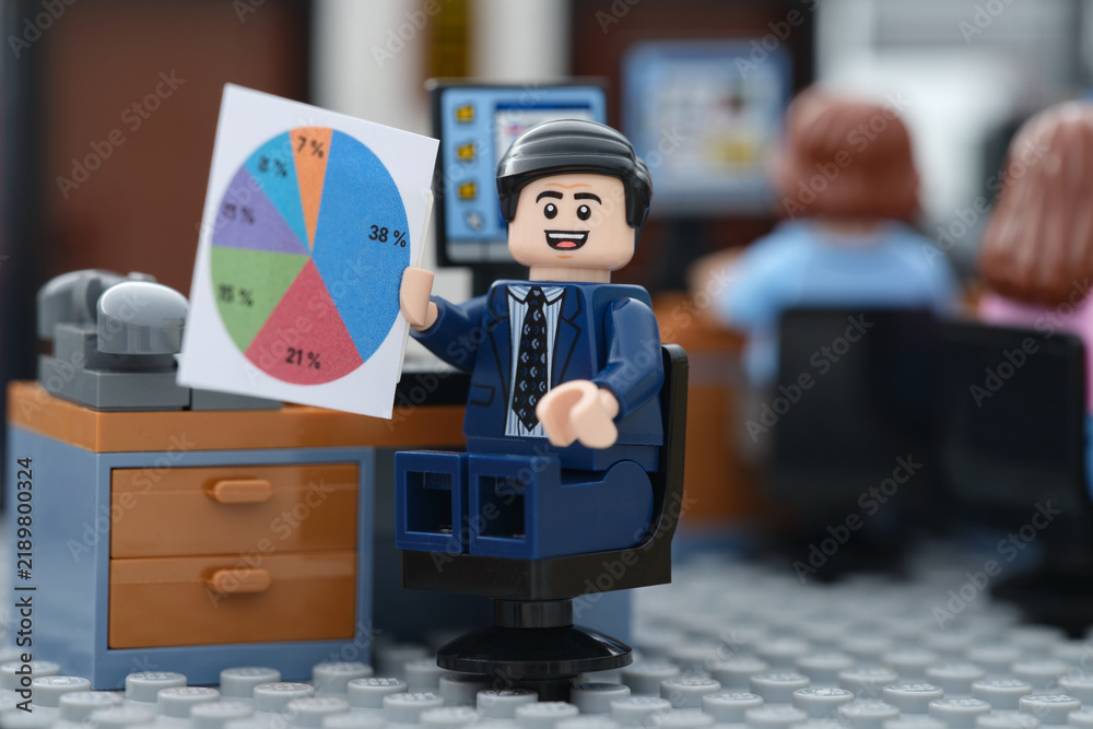 Fototapeta premium Tambov, Russian Federation - September 12, 2026 A happy Lego businessman minifigure sitting at his office desk, smiling and presenting a colorful pie chart document during a corporate meeting.