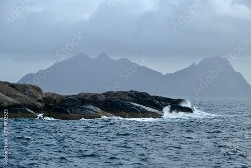 Rocky coastline with waves crashing against stones, distant islands visible under a cloudy sky in Lofoten Islands, Norway during the month of September