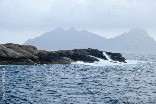 Rocky coastline with waves crashing against stones, distant islands visible under a cloudy sky in Lofoten Islands, Norway during the month of September
