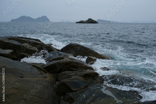 Rocky coastline with waves crashing against stones, distant islands visible under a cloudy sky in Lofoten Islands, Norway during the month of September